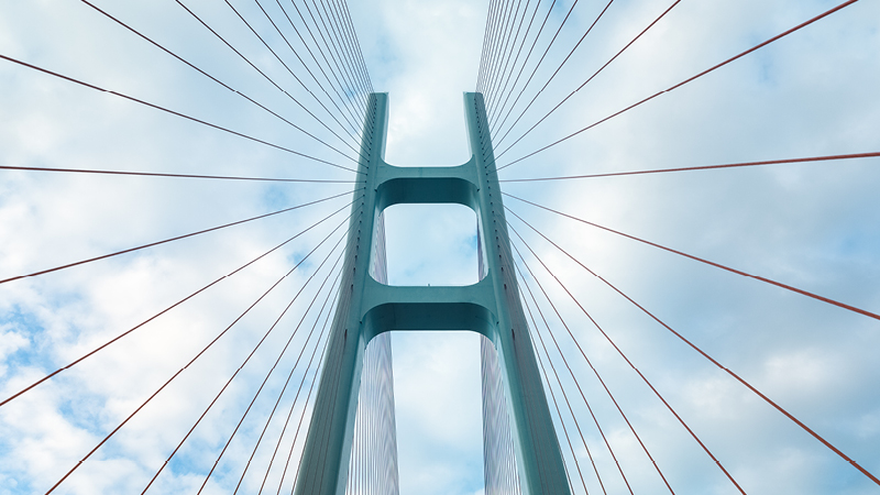 A bridge tower and cables viewed from below against a cloudy sky